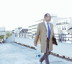 Mature businessman with skateboard and briefcase walking on parking deck - UUF19723