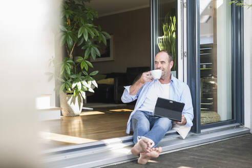 Mature man sitting at terrace door at home with tablet and coffee cup - UUF19730