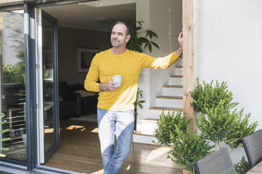 Mature man standing at terrace door at home with coffee cup - UUF19731