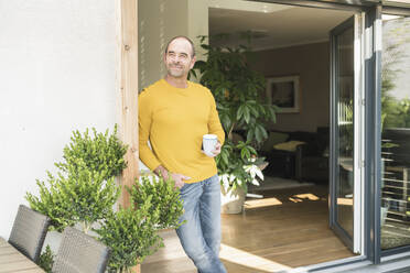 Mature man standing at terrace door at home with coffee cup - UUF19732
