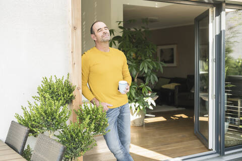 Reifer Mann mit geschlossenen Augen an der Terrassentür zu Hause mit Kaffeetasse stehend, lizenzfreies Stockfoto