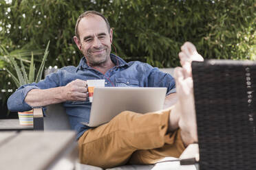 Smiling mature man sitting on terrace with earphones and laptop - UUF19734