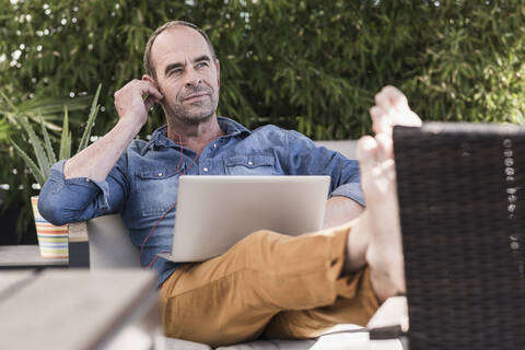 Älterer Mann sitzt mit Kopfhörern und Laptop auf der Terrasse, lizenzfreies Stockfoto