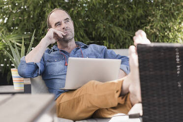 Thoughtful mature man sitting on terrace with laptop - UUF19736