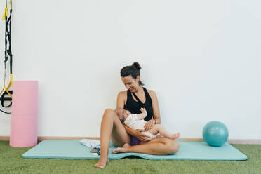 Young mother sitting on yoga mat, breastfeeding her baby - MPPF00286