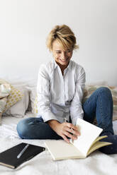 Happy mature woman with book sitting on bed at home - VABF02331