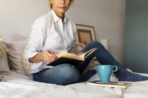 Mature woman with book and coffee cup sitting on bed at home stock photo