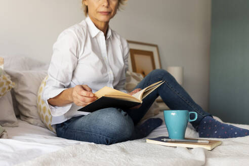 Mature woman with book and coffee cup sitting on bed at home - VABF02332