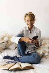 Porträt einer lächelnden reifen Frau, die zu Hause auf dem Bett sitzt und ein Smartphone hält - VABF02337
