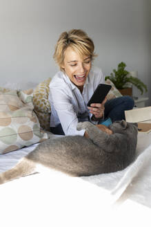 Glückliche reife Frau, die ein Smartphone-Foto von ihrer Katze auf dem Bett zu Hause macht - VABF02344