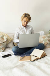 Mature woman sitting on bed at home using laptop - VABF02345