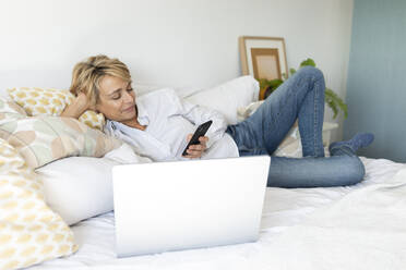 Reife Frau liegt zu Hause auf dem Bett und benutzt Smartphone und Laptop - VABF02353