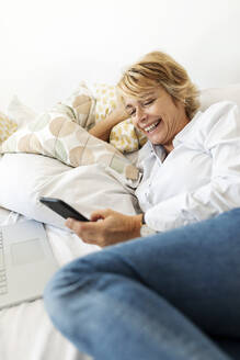 Happy mature woman sitting on bed at home using smartphone - VABF02355