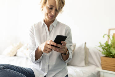 Mature woman sitting on bed at home using smartphone - VABF02357