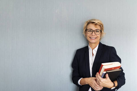 Porträt einer reifen Geschäftsfrau, die an einer Wand steht und einen Bücherstapel hält, lizenzfreies Stockfoto