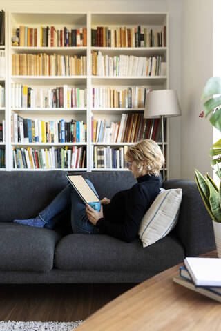 Reife Frau entspannt sich zu Hause auf der Couch mit einem Grafiktablett, lizenzfreies Stockfoto
