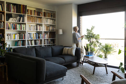 Reife Frau steht am Fenster im Wohnzimmer zu Hause, lizenzfreies Stockfoto