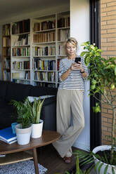 Reife Frau steht am Fenster im Wohnzimmer zu Hause und benutzt ein Smartphone - VABF02400