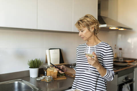 Mature woman drinking wine and using her smartphone in kitchen at home stock photo