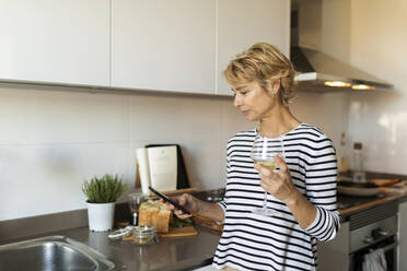 Reife Frau trinkt Wein und benutzt ihr Smartphone in der Küche zu Hause - VABF02436