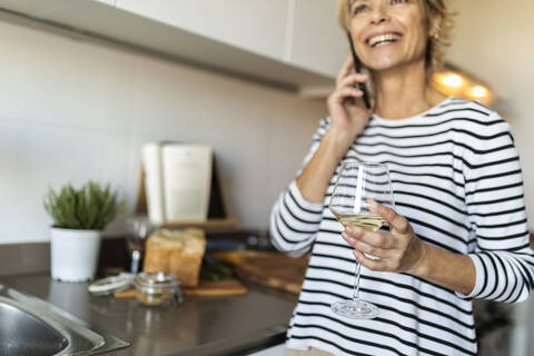 Happy mature woman drinking wine and talking on the phone in kitchen at home stock photo