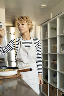 Ältere Frau, die ein hausgemachtes Nudelgericht in der Küche zu Hause probiert - VABF02454