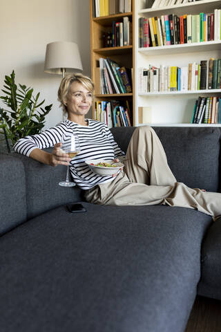 Reife Frau isst hausgemachte Nudeln und trinkt Wein auf der Couch zu Hause, lizenzfreies Stockfoto