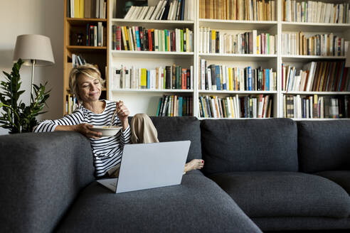 Mature woman using laptop and having lunch on couch at home - VABF02461