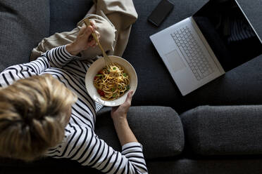Mature woman with laptop eating homemade pasta dish on couch at home - VABF02462