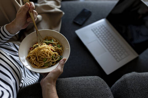 Mature woman with laptop eating homemade pasta dish on couch at home stock photo