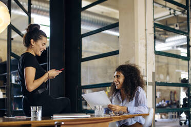 Zwei glückliche junge Geschäftsfrauen mit Smartphone im Loftbüro - SODF00360