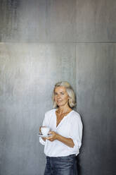 Porträt einer lässigen reifen Geschäftsfrau, die vor einer grauen Wand steht und eine Kaffeetasse hält - SODF00385