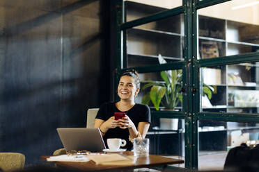 Fröhliche, lässige junge Geschäftsfrau mit Laptop und Smartphone im Loftbüro - SODF00392