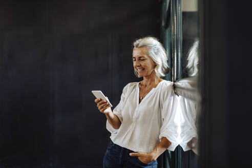 Lächelnde lässige reife Geschäftsfrau mit Smartphone am Fenster im Loft-Büro - SODF00395