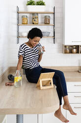 Junge Frau benutzt Tablet und trinkt Wasser in der Küche zu Hause - GIOF07782