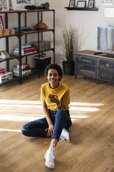 Portrait of smiling young woman sitting on the floor at home holding smartphone - GIOF07788