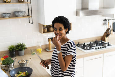 Porträt einer lächelnden jungen Frau, die zu Hause in der Küche kocht und Gemüse probiert - GIOF07791