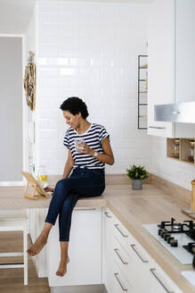 Young woman using tablet and drinking water in kitchen at home - GIOF07807