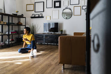 Young woman sitting on the floor at home holding smartphone - GIOF07828