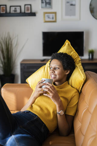 Entspannte junge Frau liegt zu Hause auf der Couch und hält eine Tasse, lizenzfreies Stockfoto