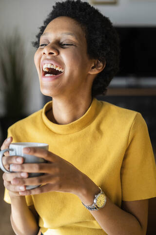 Lachende junge Frau hält Tasse zu Hause, lizenzfreies Stockfoto