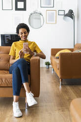 Smiling young woman sitting on couch at home using smartphone - GIOF07837