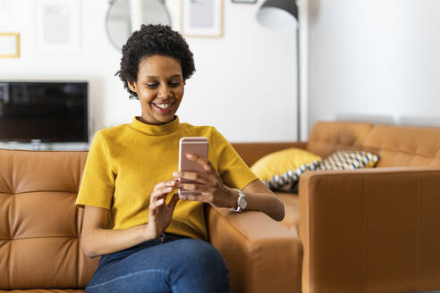 Smiling young woman sitting on couch at home using smartphone - GIOF07838