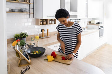 Young woman with tablet cooking in kitchen at home cutting vegetables - GIOF07842