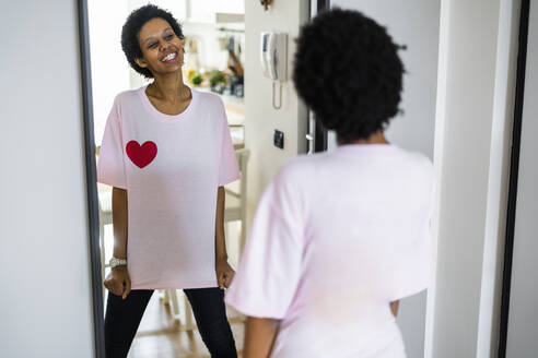 Glückliche junge Frau trägt T-Shirt mit Herzform und schaut in den Spiegel - GIOF07857