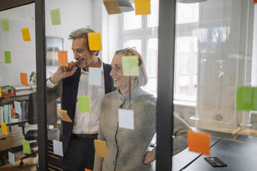 Glückliche Kollegen, die auf Haftnotizen an einer Glasscheibe im Büro schauen - GUSF02682