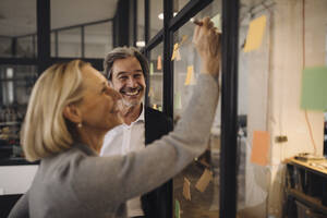 Happy colleagues with sticky notes at glass pane in office - GUSF02684