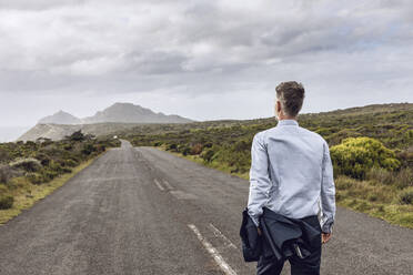 Rückenansicht eines Geschäftsmannes, der auf einer Landstraße spazieren geht, Cape Point, Westkap, Südafrika - MCF00337