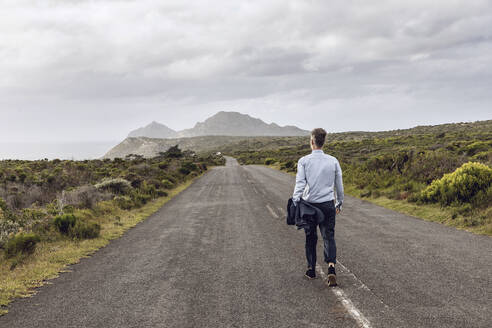 Rückenansicht eines Geschäftsmannes, der auf einer Landstraße spazieren geht, Cape Point, Westkap, Südafrika - MCF00340