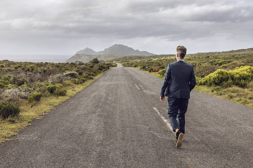 Rückenansicht eines Geschäftsmannes, der auf einer Landstraße spazieren geht, Cape Point, Westkap, Südafrika - MCF00341